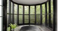 Curved wall of black shutters with greenery visible outside and a small circular tiled hot tub in a modern spa room.