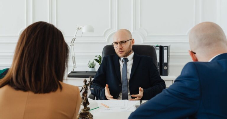 Economic abuse - Beanstalk Mums A bald man in a suit speaks to two colleagues in an office, gesturing with hands over a desk with documents and a statue centerpiece