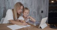 Mother helps her young son with homework at a wooden table, writing in a notebook with a pencil beside a tablet and books.