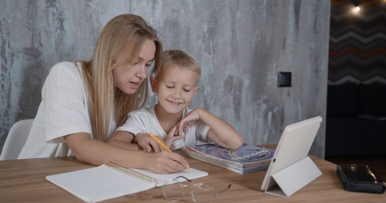 Mother helps her young son with homework at a wooden table, writing in a notebook with a pencil beside a tablet and books.