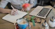 Person writes in a lined notebook at a wooden desk next to a laptop and a cup of coffee, with a blurred flower in the foreground.