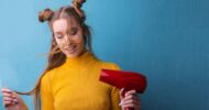 Woman with two buns and glasses uses a red hair dryer against a blue wall.