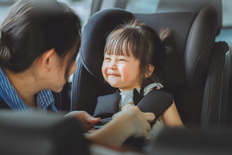 Image - Beanstalk Mums Young girl smiling while being buckled into a car seat by a caregiver in a car interior