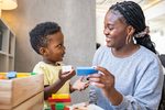 Young child and woman smiling while stacking colorful wooden blocks at a table in a bright play area.