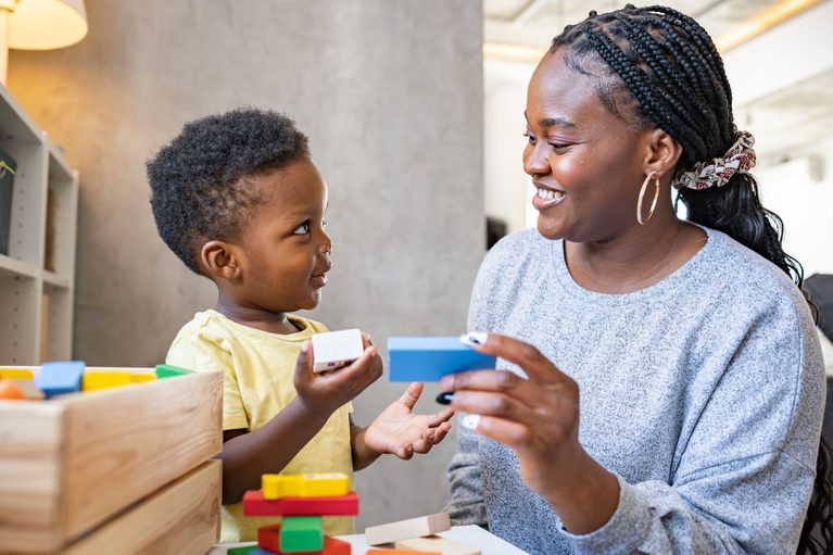 Young child and woman smiling while stacking colorful wooden blocks at a table in a bright play area.